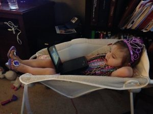 Three year old girl laying in a baby rocker with princess gear and a DVD player, with tax books on the bookshelf behind her
